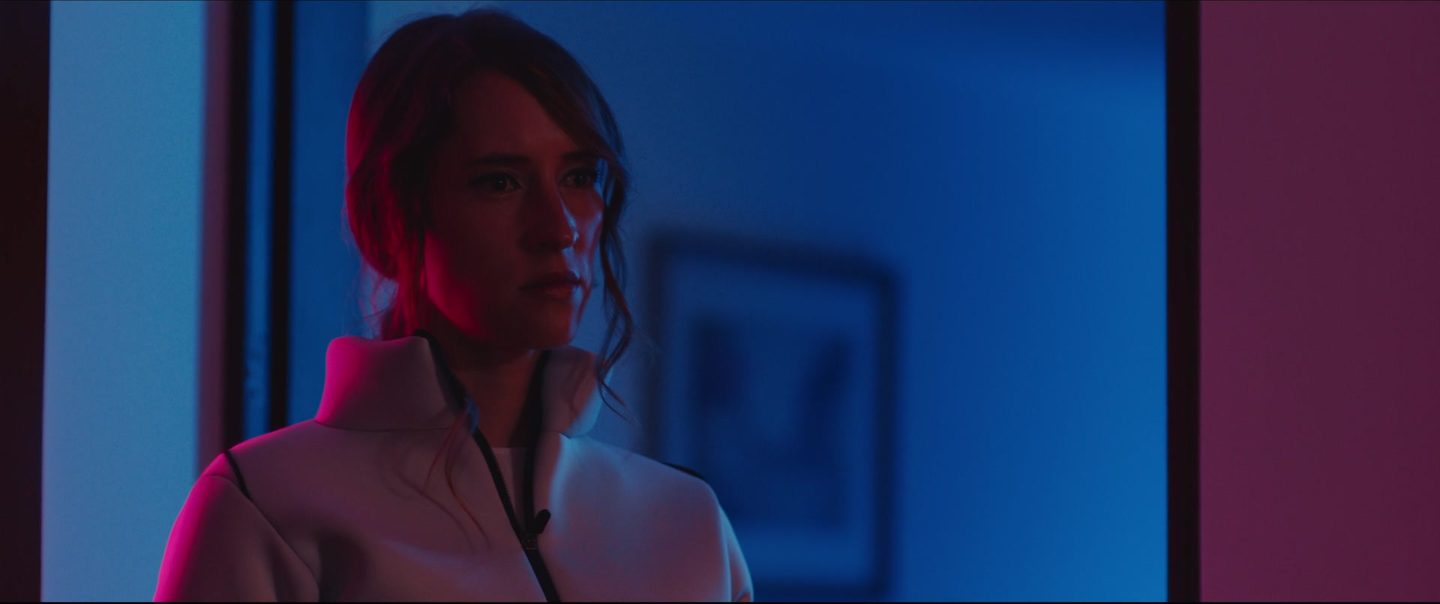Still from Infinite Regress: a woman in a white jacket in a room lit red and blue